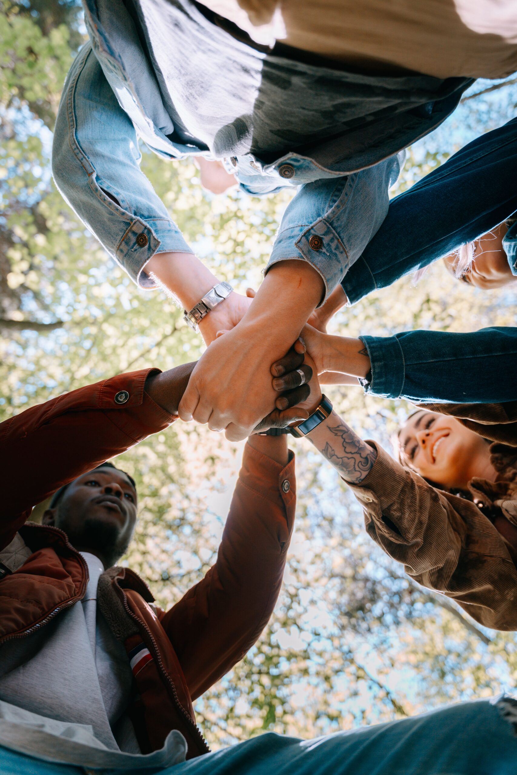 A diverse group of adults joining hands outdoors, symbolizing unity and teamwork from a low angle perspective.